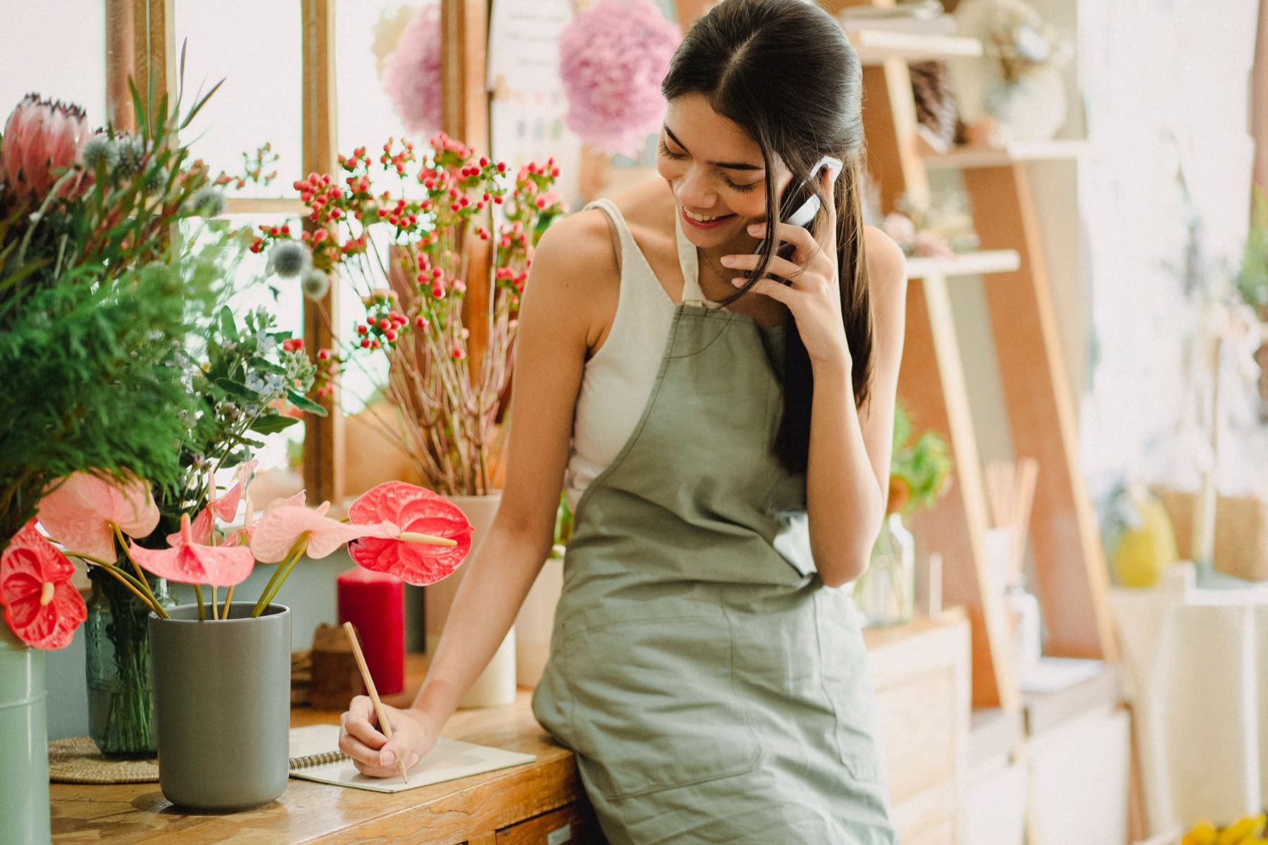 A shop owner in an apron smiles while talking on her phone at her work table.